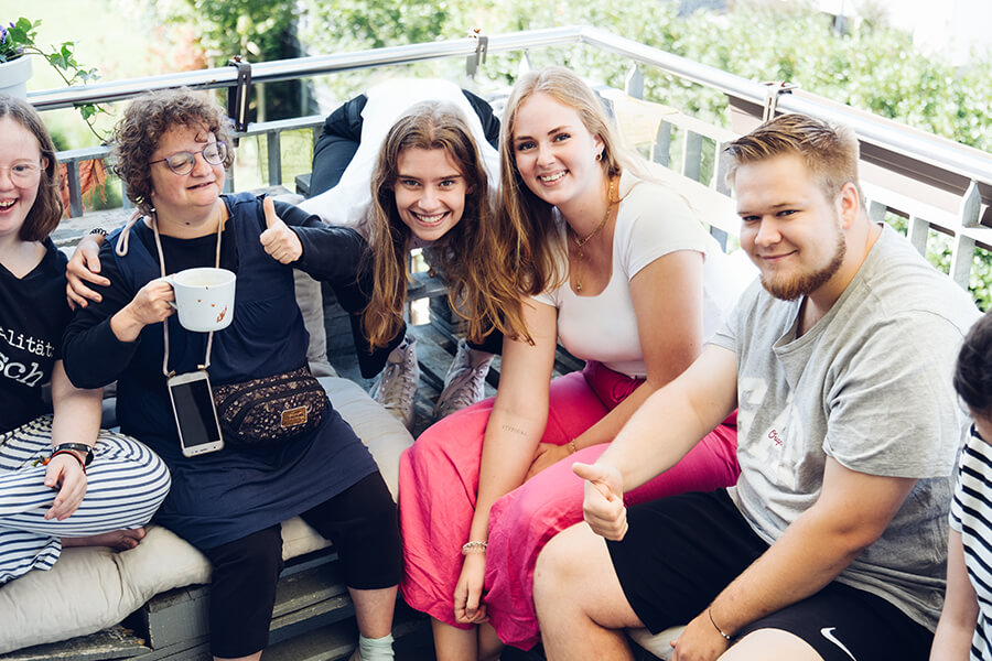 Vier junge Bewohner einer inklusiven WG sitzen auf der Terrasse. Alle lächeln freundlich in die Kamera.